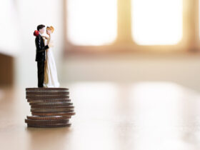 Casal de noivos em miniatura sobre uma pilha de moedas simbolizando planejamento financeiro e como juntar dinheiro para o casamento de forma estratégica.