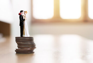 Casal de noivos em miniatura sobre uma pilha de moedas simbolizando planejamento financeiro e como juntar dinheiro para o casamento de forma estratégica.