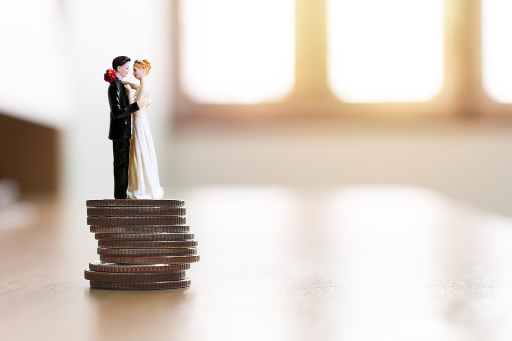 Casal de noivos em miniatura sobre uma pilha de moedas simbolizando planejamento financeiro e como juntar dinheiro para o casamento de forma estratégica.