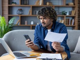 Como quitar dívidas: passo a passo para limpar seu nome definitivamente 4 Um homem jovem de cabelos cacheados e barba curta está sentado em um sofá cinza, segurando um boleto em uma das mãos enquanto consulta o celular com a outra. Ele apresenta uma expressão de concentração e preocupação. À sua frente, sobre uma mesa de centro de madeira, há um notebook aberto, mais papéis e uma caneta, ilustrando o momento de organização financeira sobre como quitar dívidas de forma planejada no ambiente doméstico.