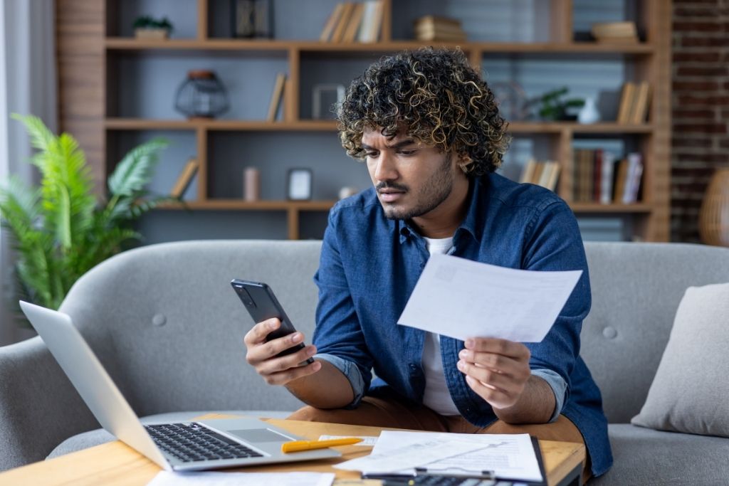 Como quitar dívidas: passo a passo para limpar seu nome definitivamente 1 Um homem jovem de cabelos cacheados e barba curta está sentado em um sofá cinza, segurando um boleto em uma das mãos enquanto consulta o celular com a outra. Ele apresenta uma expressão de concentração e preocupação. À sua frente, sobre uma mesa de centro de madeira, há um notebook aberto, mais papéis e uma caneta, ilustrando o momento de organização financeira sobre como quitar dívidas de forma planejada no ambiente doméstico.