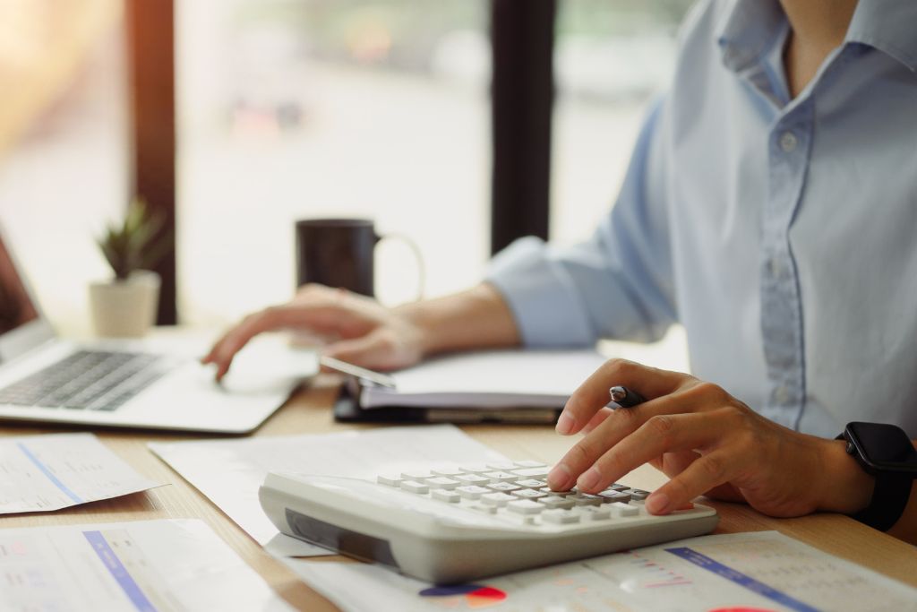 Pessoa usando calculadora e notebook ao mesmo tempo, organizando dados financeiros em uma mesa de trabalho, representando a rotina prática de controle de recebimento no dia a dia de pequenos negócios.