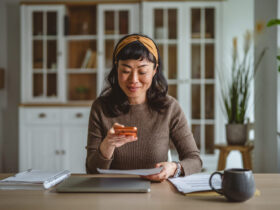 Fluxo de caixa pessoal: o segredo para nunca ficar no vermelho antes do dia 15 4 Uma mulher de ascendência asiática com cabelos escuros e uma tiara amarela está sentada à mesa de sua casa organizada. Ela demonstra foco ao utilizar o celular para fotografar ou digitalizar um documento impresso, possivelmente uma conta ou comprovante para atualizar seu fluxo de caixa pessoal. Sobre a mesa de madeira clara, há um notebook fechado, um caderno de espiral, uma caneta e uma caneca cinza, criando um ambiente doméstico ideal para a gestão financeira. Ao fundo, vê-se um armário branco com portas de vidro e uma planta decorativa, reforçando a sensação de ordem e cuidado com as finanças.