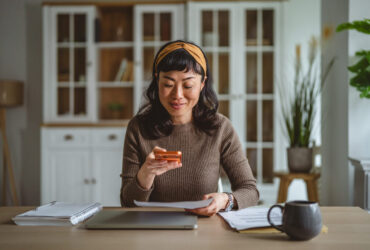 Uma mulher de ascendência asiática com cabelos escuros e uma tiara amarela está sentada à mesa de sua casa organizada. Ela demonstra foco ao utilizar o celular para fotografar ou digitalizar um documento impresso, possivelmente uma conta ou comprovante para atualizar seu fluxo de caixa pessoal. Sobre a mesa de madeira clara, há um notebook fechado, um caderno de espiral, uma caneta e uma caneca cinza, criando um ambiente doméstico ideal para a gestão financeira. Ao fundo, vê-se um armário branco com portas de vidro e uma planta decorativa, reforçando a sensação de ordem e cuidado com as finanças.