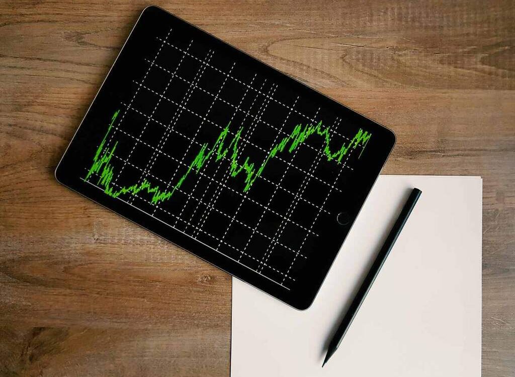 Tablet exibindo gráfico financeiro com linhas verdes sobre fundo preto, indicando variações de mercado ao longo do tempo. Ao lado, uma folha em branco e uma caneta preta repousam sobre uma superfície de madeira, sugerindo um ambiente de análise e planejamento. Imagem representa o compromisso dos investimentos sustentáveis com decisões baseadas em dados e responsabilidade financeira.