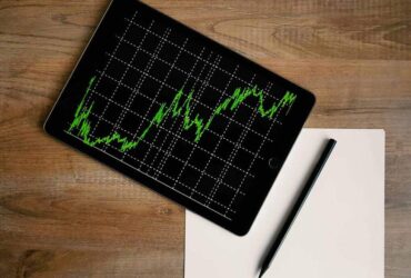 Tablet exibindo gráfico financeiro com linhas verdes sobre fundo preto, indicando variações de mercado ao longo do tempo. Ao lado, uma folha em branco e uma caneta preta repousam sobre uma superfície de madeira, sugerindo um ambiente de análise e planejamento. Imagem representa o compromisso dos investimentos sustentáveis com decisões baseadas em dados e responsabilidade financeira.