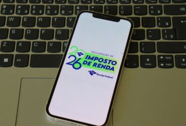Um smartphone posicionado sobre o teclado de um laptop exibe em sua tela o logotipo oficial da Receita Federal para a declaração de IR 2026. A imagem ilustra a facilidade de acesso digital para acompanhar a situação da declaração e verificar se o contribuinte apresenta pendências que possam levá-lo para a malha fina do Imposto de Renda.