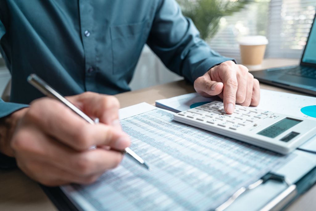 Fotografia mostra em detalhe as mãos de uma pessoa utilizando uma calculadora e uma caneta para conferir dados em planilhas financeiras impressas. A cena representa o cuidado e a revisão minuciosa necessária durante a organização de documentos para evitar inconsistências nos dados enviados e prevenir problemas com a malha fina do Imposto de Renda.