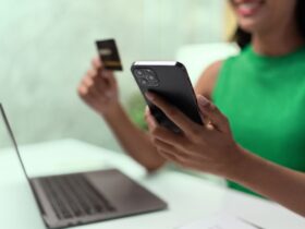 Mulher vestindo verde, cor da Organizze, sorrindo enquanto utiliza o celular e segura um cartão de crédito em frente a um notebook, representando o momento de escolha consciente e o uso de tecnologia para controle financeiro.
