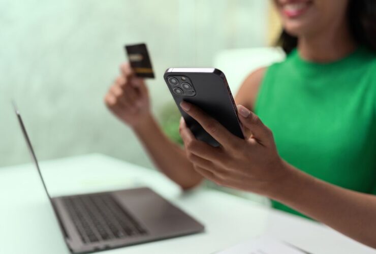 Mulher vestindo verde, cor da Organizze, sorrindo enquanto utiliza o celular e segura um cartão de crédito em frente a um notebook, representando o momento de escolha consciente e o uso de tecnologia para controle financeiro.