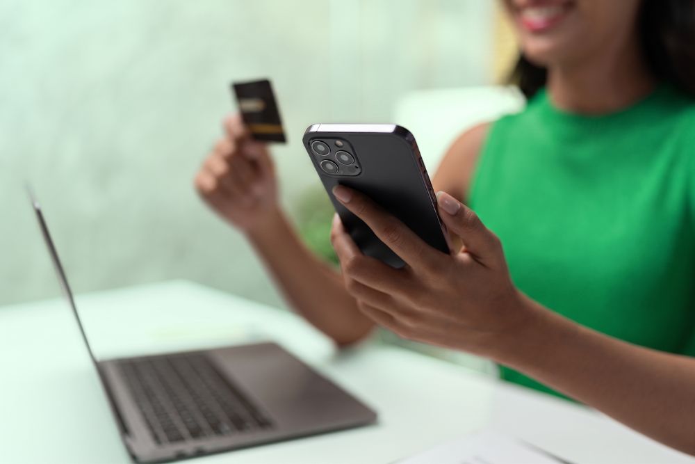 Mindfulness financeiro: como a atenção plena transforma sua relação com o dinheiro 1 Mulher vestindo verde, cor da Organizze, sorrindo enquanto utiliza o celular e segura um cartão de crédito em frente a um notebook, representando o momento de escolha consciente e o uso de tecnologia para controle financeiro.