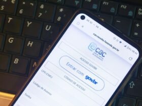 Uma foto em close mostra a tela de um smartphone posicionado sobre o teclado de um notebook. O aparelho exibe a página de login do portal da Receita Federal, destacando o logotipo do Centro Virtual de Atendimento ao Contribuinte. Abaixo do logo, visualizam-se as opções de acesso por meio da conta gov.br ou por código de acesso com CPF e CNPJ. A imagem ilustra de forma prática o que é o e-CAC e como a interface do sistema aparece para o usuário no ambiente mobile.
