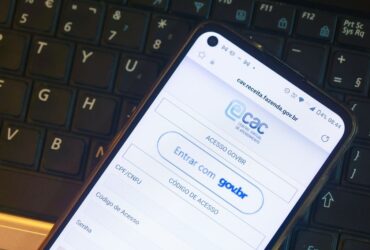 Uma foto em close mostra a tela de um smartphone posicionado sobre o teclado de um notebook. O aparelho exibe a página de login do portal da Receita Federal, destacando o logotipo do Centro Virtual de Atendimento ao Contribuinte. Abaixo do logo, visualizam-se as opções de acesso por meio da conta gov.br ou por código de acesso com CPF e CNPJ. A imagem ilustra de forma prática o que é o e-CAC e como a interface do sistema aparece para o usuário no ambiente mobile.