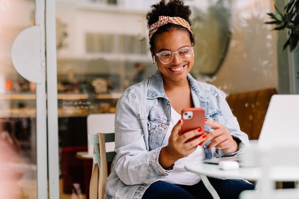 O que é e-CAC e por que você precisa conhecer o portal da Receita 3 Uma mulher jovem e sorridente, com cabelos presos em um coque e usando óculos, está sentada em uma mesa de café ao ar livre enquanto utiliza o celular. Ela veste uma jaqueta jeans e parece tranquila ao navegar no aparelho, que segura com as duas mãos. Sobre a mesa, há um notebook e fones de ouvido sem fio. A cena transmite a facilidade de realizar consultas fiscais e entender o que é o e-CAC de qualquer lugar, aproveitando a mobilidade dos serviços digitais do governo.