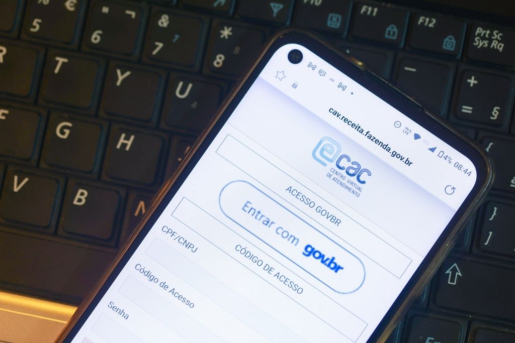 O que é e-CAC e por que você precisa conhecer o portal da Receita 1 Uma foto em close mostra a tela de um smartphone posicionado sobre o teclado de um notebook. O aparelho exibe a página de login do portal da Receita Federal, destacando o logotipo do Centro Virtual de Atendimento ao Contribuinte. Abaixo do logo, visualizam-se as opções de acesso por meio da conta gov.br ou por código de acesso com CPF e CNPJ. A imagem ilustra de forma prática o que é o e-CAC e como a interface do sistema aparece para o usuário no ambiente mobile.