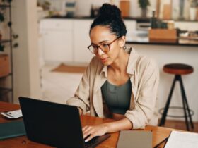 Uma mulher jovem de pele parda com o cabelo preso em um coque alto e óculos de armação fina está sentada à mesa de madeira em um ambiente doméstico iluminado. Ela demonstra concentração enquanto utiliza um notebook preto para gerenciar suas finanças pessoais. Sobre a mesa além do computador estão um caderno cinza fechado e uma calculadora branca, reforçando a ideia de planejamento detalhado necessário para aplicar o orçamento base zero. O ambiente ao fundo mostra uma cozinha moderna levemente desfocada sugerindo um contexto de organização do dia a dia familiar.
