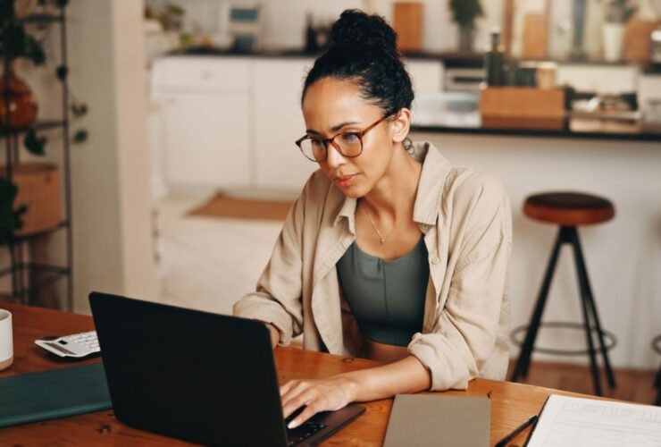 Uma mulher jovem de pele parda com o cabelo preso em um coque alto e óculos de armação fina está sentada à mesa de madeira em um ambiente doméstico iluminado. Ela demonstra concentração enquanto utiliza um notebook preto para gerenciar suas finanças pessoais. Sobre a mesa além do computador estão um caderno cinza fechado e uma calculadora branca, reforçando a ideia de planejamento detalhado necessário para aplicar o orçamento base zero. O ambiente ao fundo mostra uma cozinha moderna levemente desfocada sugerindo um contexto de organização do dia a dia familiar.