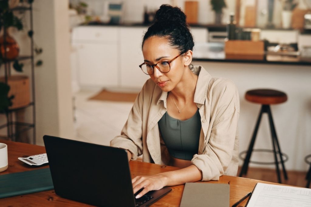 Uma mulher jovem de pele parda com o cabelo preso em um coque alto e óculos de armação fina está sentada à mesa de madeira em um ambiente doméstico iluminado. Ela demonstra concentração enquanto utiliza um notebook preto para gerenciar suas finanças pessoais. Sobre a mesa além do computador estão um caderno cinza fechado e uma calculadora branca, reforçando a ideia de planejamento detalhado necessário para aplicar o orçamento base zero. O ambiente ao fundo mostra uma cozinha moderna levemente desfocada sugerindo um contexto de organização do dia a dia familiar.