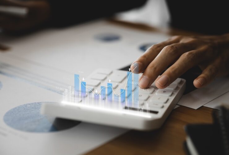 Mão digitando em calculadora com gráficos flutuantes, representando análise de dados e planejamento tributário empresarial.