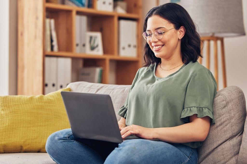 Uma mulher de óculos e blusa verde sorri enquanto utiliza um notebook sentada confortavelmente em um sofá cinza. O ambiente doméstico é acolhedor, com uma estante de livros ao fundo, sugerindo um momento de tranquilidade e autonomia. Ela demonstra satisfação ao pesquisar as melhores opções de ativos financeiros para alocar sua reserva de emergência com segurança e rentabilidade.