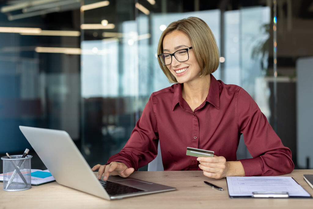 Mulher sorridente usando notebook e segurando cartão de crédito em ambiente de escritório